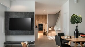 Een moderne woonkamer met een flatscreen televisie aan een donkere muur, een eethoek met een ronde houten tafel en zwarte stoelen, en een keuken op de achtergrond met houten kastjes en een inbouwoven.