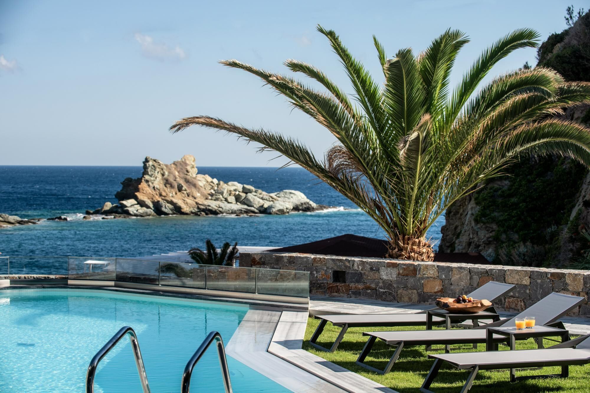 A poolside area with lounge chairs and a palm tree, overlooking a rocky coastline and the sea.