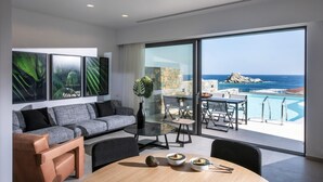 Een moderne woonkamer met een grijze bank, een glazen salontafel en een eethoek met een houten tafel en stoelen. Een groot raam biedt uitzicht op de oceaan en een rotsachtige klif.