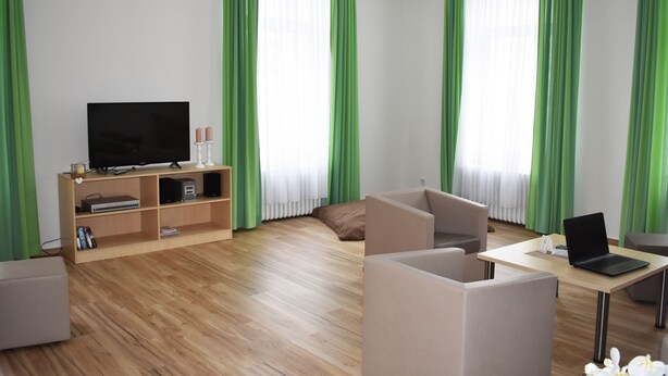 Una sala de estar moderna con un televisor de pantalla plana, un mueble de entretenimiento de madera, una computadora portátil sobre una mesa y cortinas verdes.