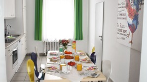 Un comedor con una mesa dispuesta para cuatro, sillas, un ventanal con cortinas verdes y una zona de cocina con armarios blancos.