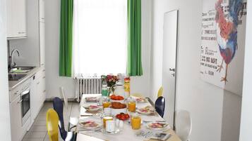 Ein Essbereich mit Tisch für vier Personen, Stühlen, einem Fenster mit grünen Vorhängen und einer Küche mit weißen Schränken.