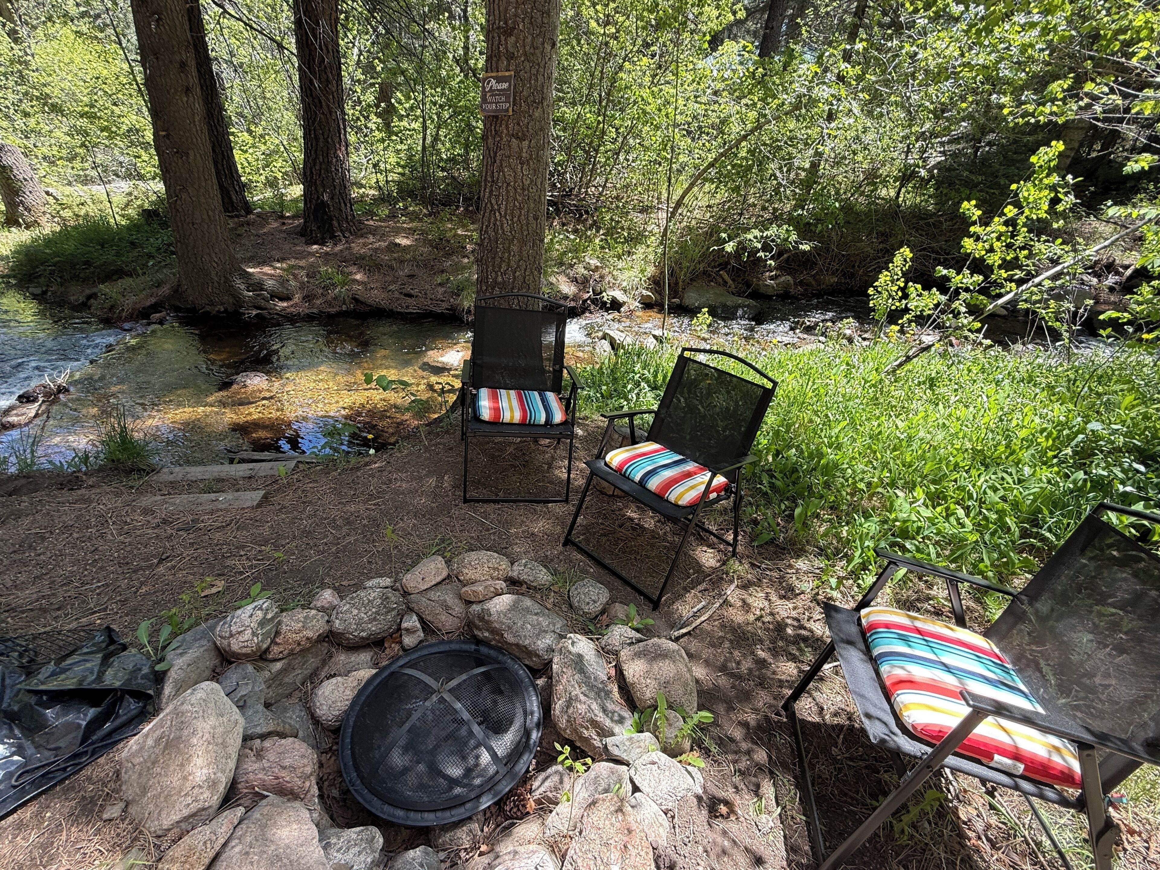 Un espacio al aire libre con dos sillas y una chimenea, rodeado de árboles y un arroyo.