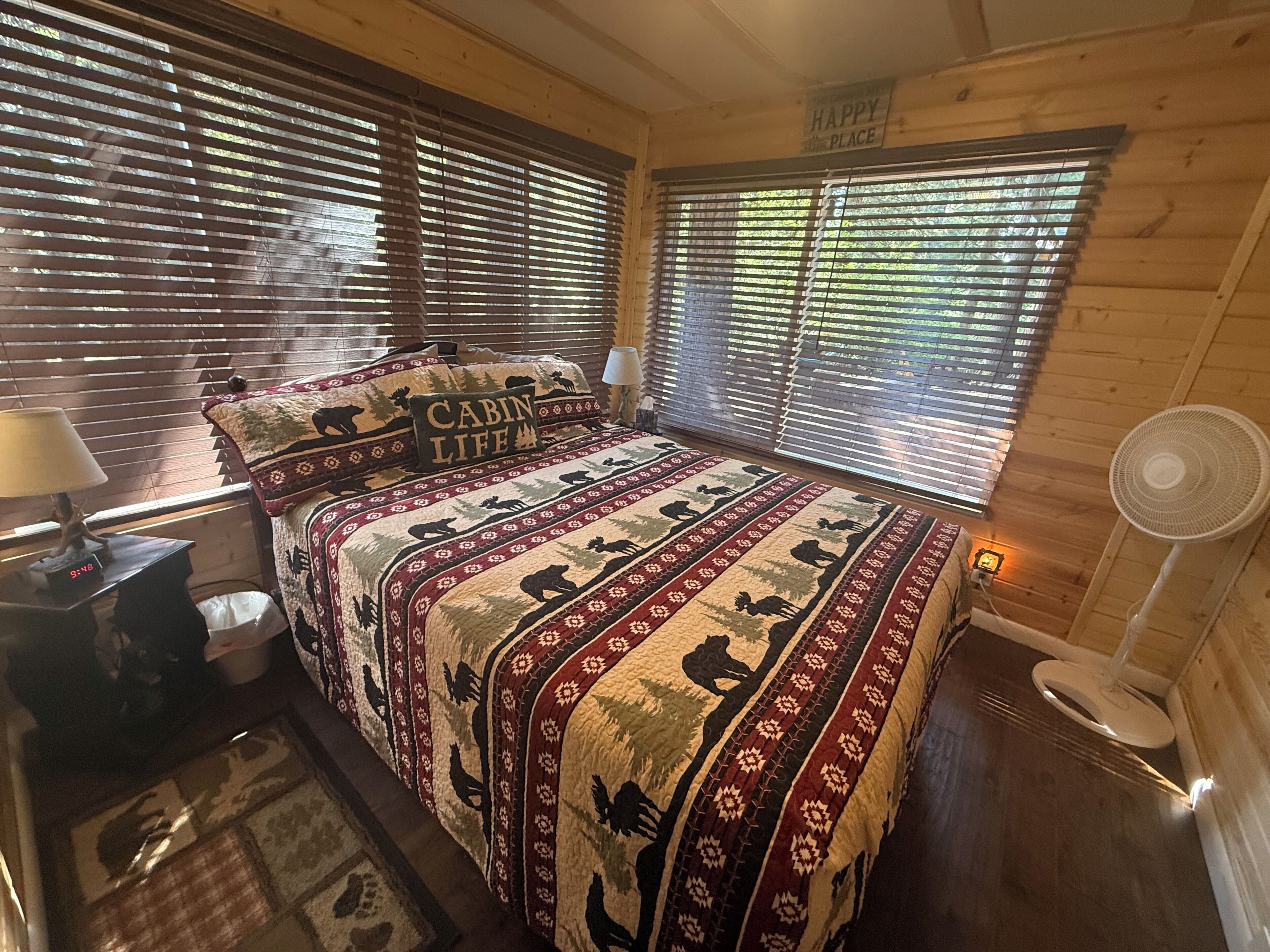 Un dormitorio tipo cabaña con una cama que tiene una colcha con motivos de animales, una lámpara de noche, un reloj y un ventilador.