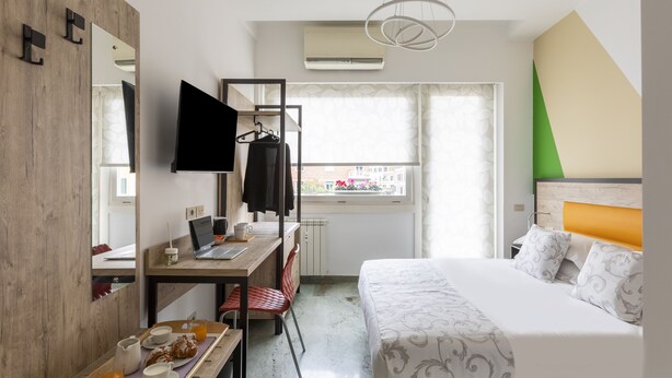 Une chambre d’hôtel avec un lit, un bureau avec un ordinateur portable, une chaise, une télévision et une fenêtre donnant sur des immeubles.