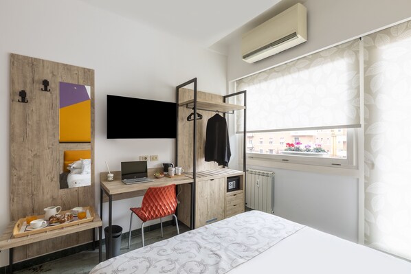 A modern hotel room with a wooden desk, a laptop, a red chair, a bed with a patterned bedspread, and a window with a view of buildings.