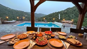 Mesa de jantar posta com diversos pratos, piscina e paisagem montanhosa ao fundo.