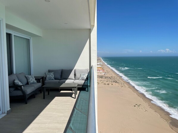 A balcony with a sofa and table, overlooking a beach with waves. - Stunning Views - 18th Floor - 1 Bedroom Deluxe (Puerto Peñasco)