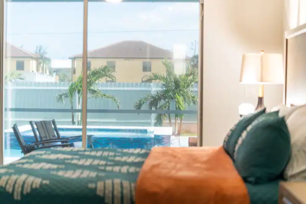 Habitación de hotel con un amplio ventanal con vistas a una piscina y palmeras.