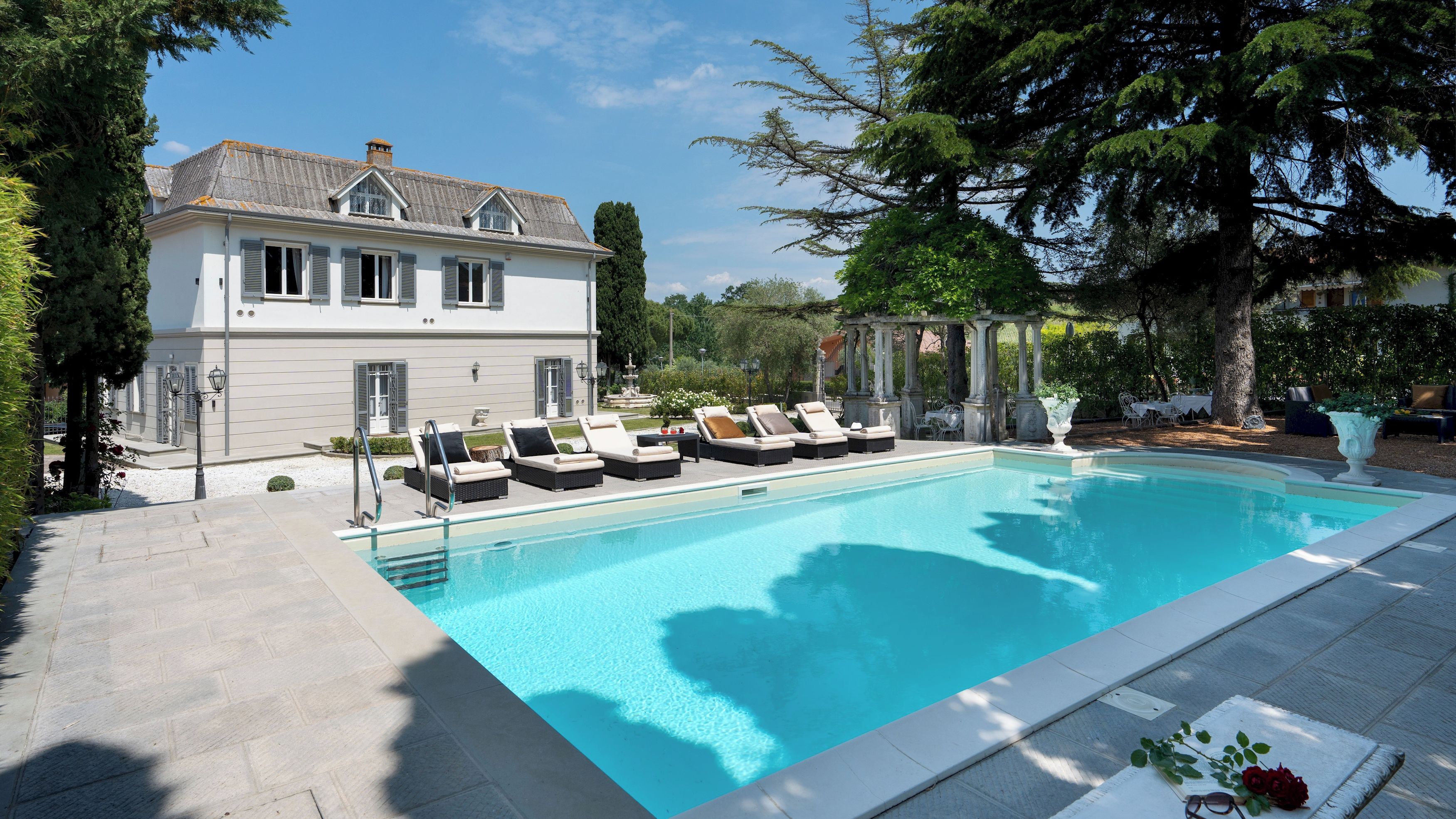 A swimming pool with lounge chairs, a white building with shutters, and a pergola with columns.