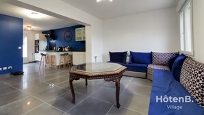Una sala de estar moderna con una mesa de centro de vidrio, un conjunto de sofás y una cocina al fondo.