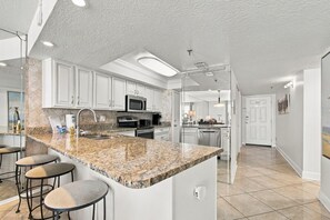 A modern kitchen with granite countertops, stainless steel appliances, and bar stools. - Beachfront Madeira Beach Condo | 1st Floor Easy Access | Heated Pool + Hot Tub | Sleeps 8 (Madeira Beach)
