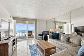 A modern living room with a large TV, leather sofa, and a balcony with a view of the beach. - Beachfront Madeira Beach Condo | 1st Floor Easy Access | Heated Pool + Hot Tub | Sleeps 8 (Madeira Beach)
