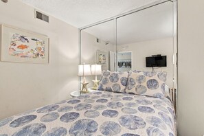 A bedroom with a bed, a bedside table with lamps, a television, and a mirror. - Beachfront Madeira Beach Condo | 1st Floor Easy Access | Heated Pool + Hot Tub | Sleeps 8 (Madeira Beach)
