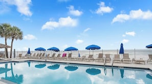 Una piscina con sillones de descanso y sombrillas, cielo despejado y una palmera.