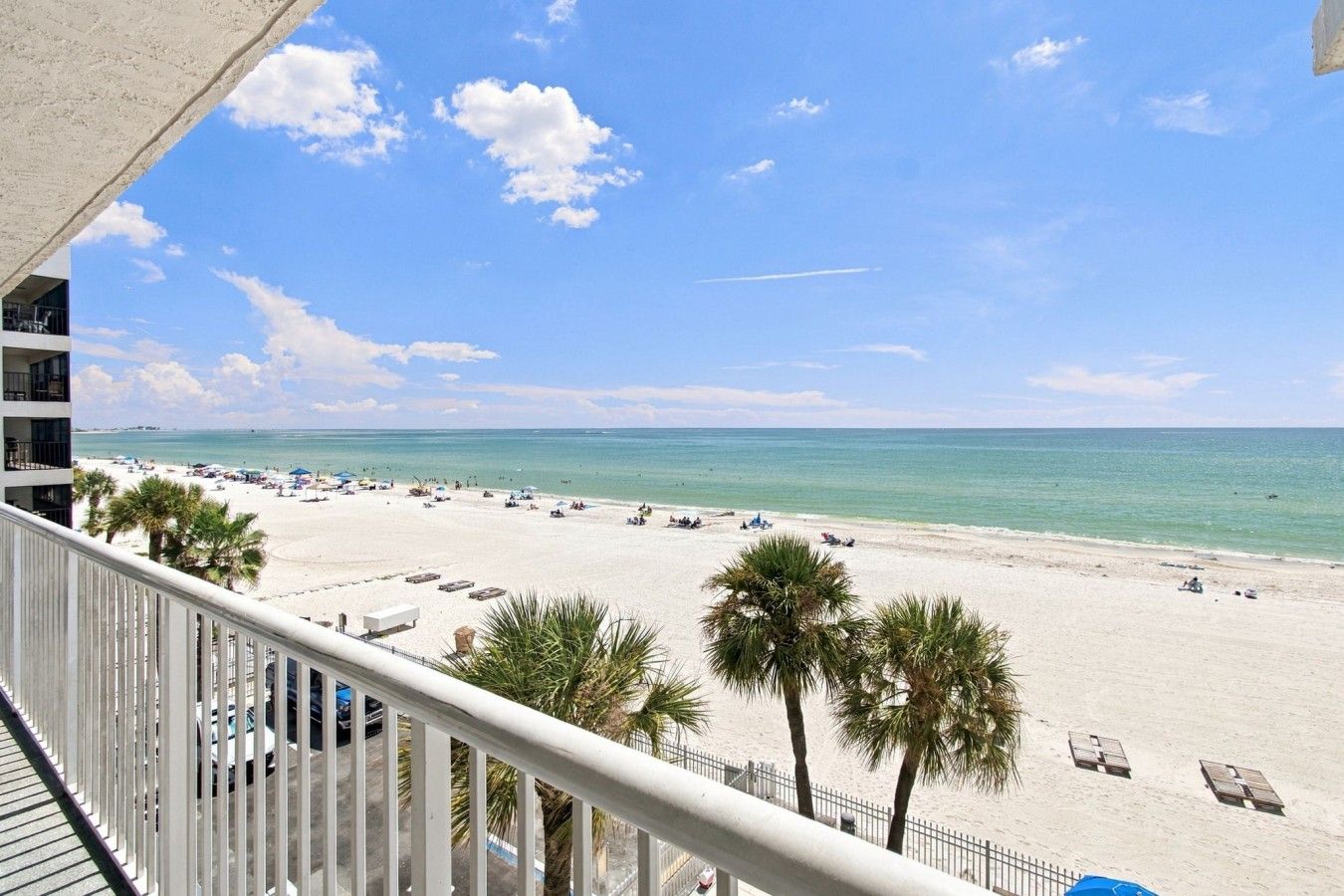 A beach view from a balcony with palm trees, a clear sky, and a sandy beach with umbrellas and lounge chairs.