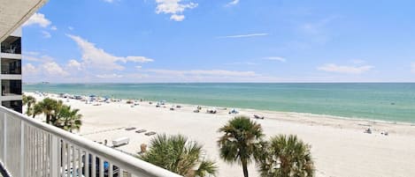 A beach view from a balcony with palm trees, a clear sky, and a sandy beach with umbrellas and lounge chairs.