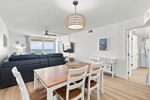 Uma sala de estar moderna com sofá, mesa de jantar, cadeiras, ventilador de teto e vista para o oceano através de amplas janelas.