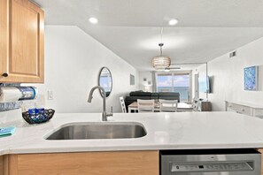 A modern kitchen with a built-in dishwasher, a large sink, wooden cabinets, and a dining area with a view of the ocean.