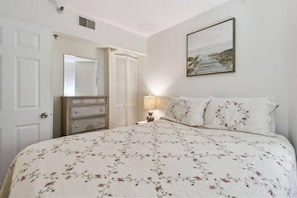 Un dormitorio con colcha estampada, lámpara de noche, espejo y un cuadro en la pared.