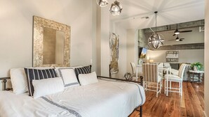 A modern bedroom with a large bed, a dining area with a table and chairs, and a kitchen in the background.