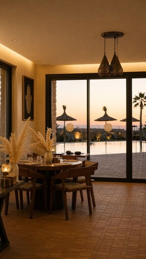 An indoor dining area with a table set for four, large windows with a view of a body of water and palm trees, and hanging pendant lights. - Baberrih Hospitality Palaces & Resorts (Moulay Bouzarqtoune)