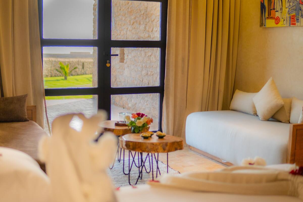 A room with a bed, a sofa, a small round table with a vase of flowers, and a view through a large window.