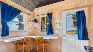 A wooden cabin interior with a white countertop, two wooden chairs, a door, and a window with blue curtains. - Quiet Glamping Barn on a Working Farm with Fire Pit and Open Views (Lynden)