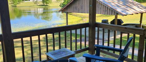 A wooden deck with Adirondack chairs and a table, overlooking a lake and a building.