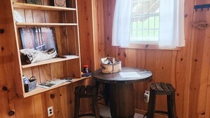 Una habitación de madera con una mesa redonda y dos taburetes, una estantería de madera con diversos objetos, y una ventana con cortinas blancas.