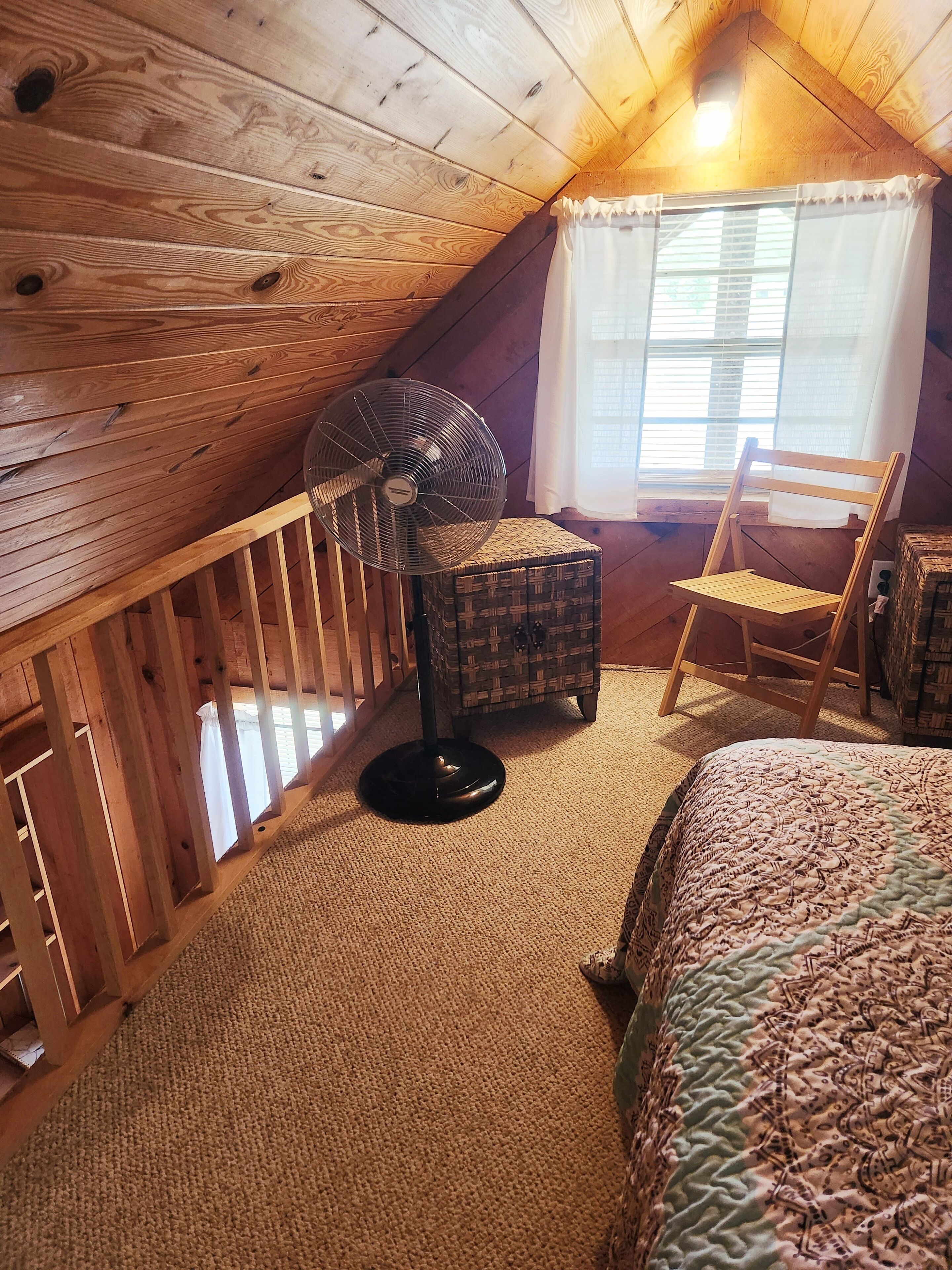 Un dormitorio acogedor en el ático, con paredes de madera y techo inclinado. Tiene una cama grande, un sillón mecedora, una mesita pequeña y un ventilador de pie.