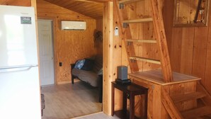 Interior de una cabaña de madera con una cama, una mesita y un refrigerador.