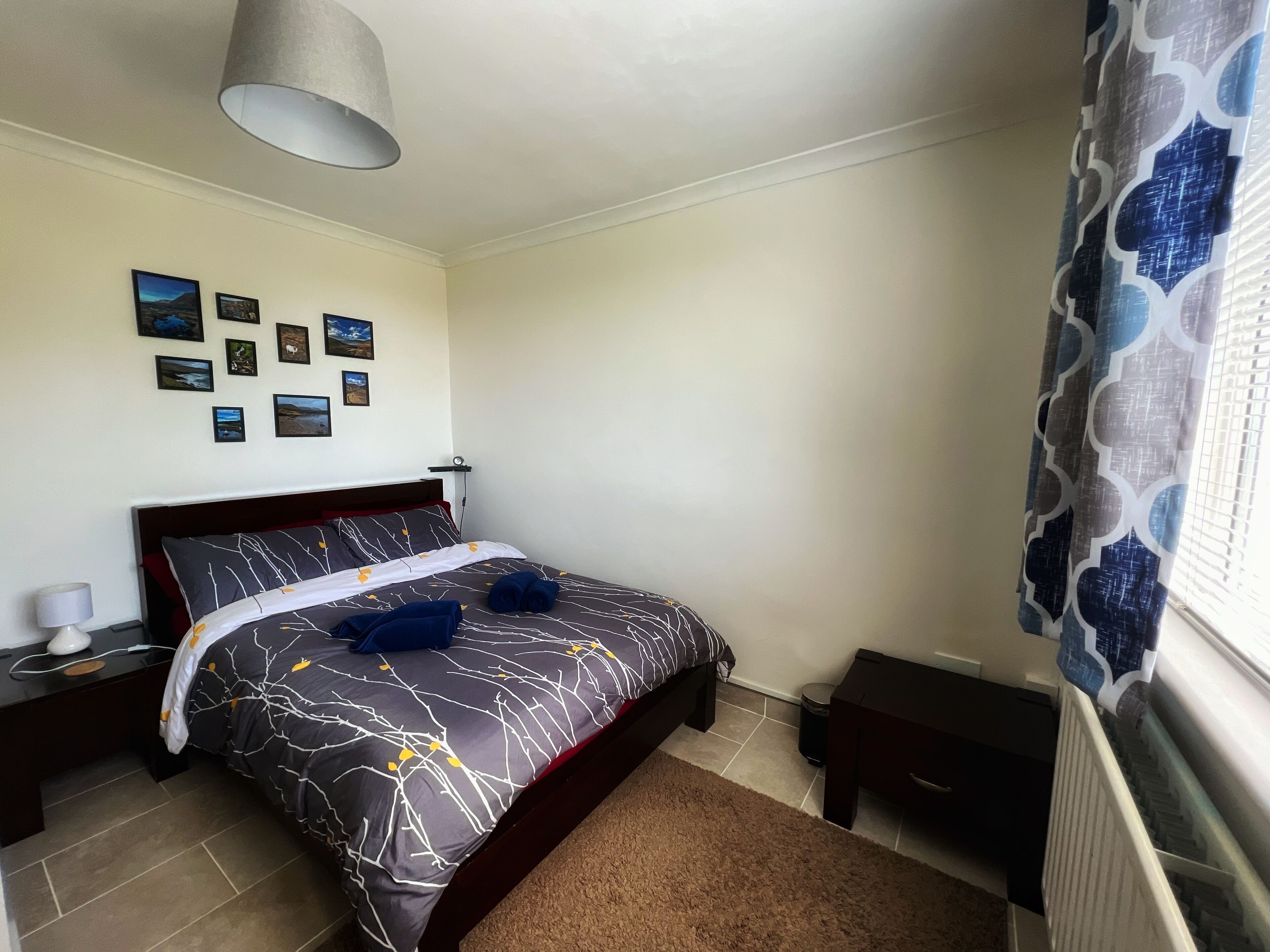 A neatly arranged bedroom with a bed, bedside tables, a lamp, and a wall with framed pictures.