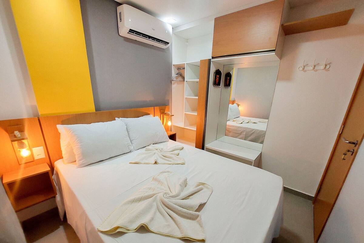 A hotel room with a bed, bedside table, nightstand, wardrobe, mirror, and an air conditioning unit.