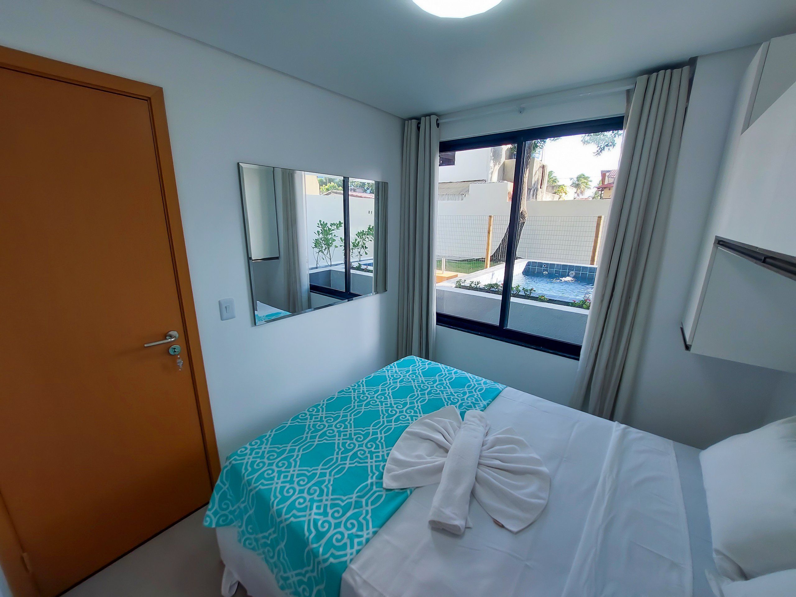 A hotel room with a bed, a window with a view of a swimming pool, a door, and a mirror on the wall.