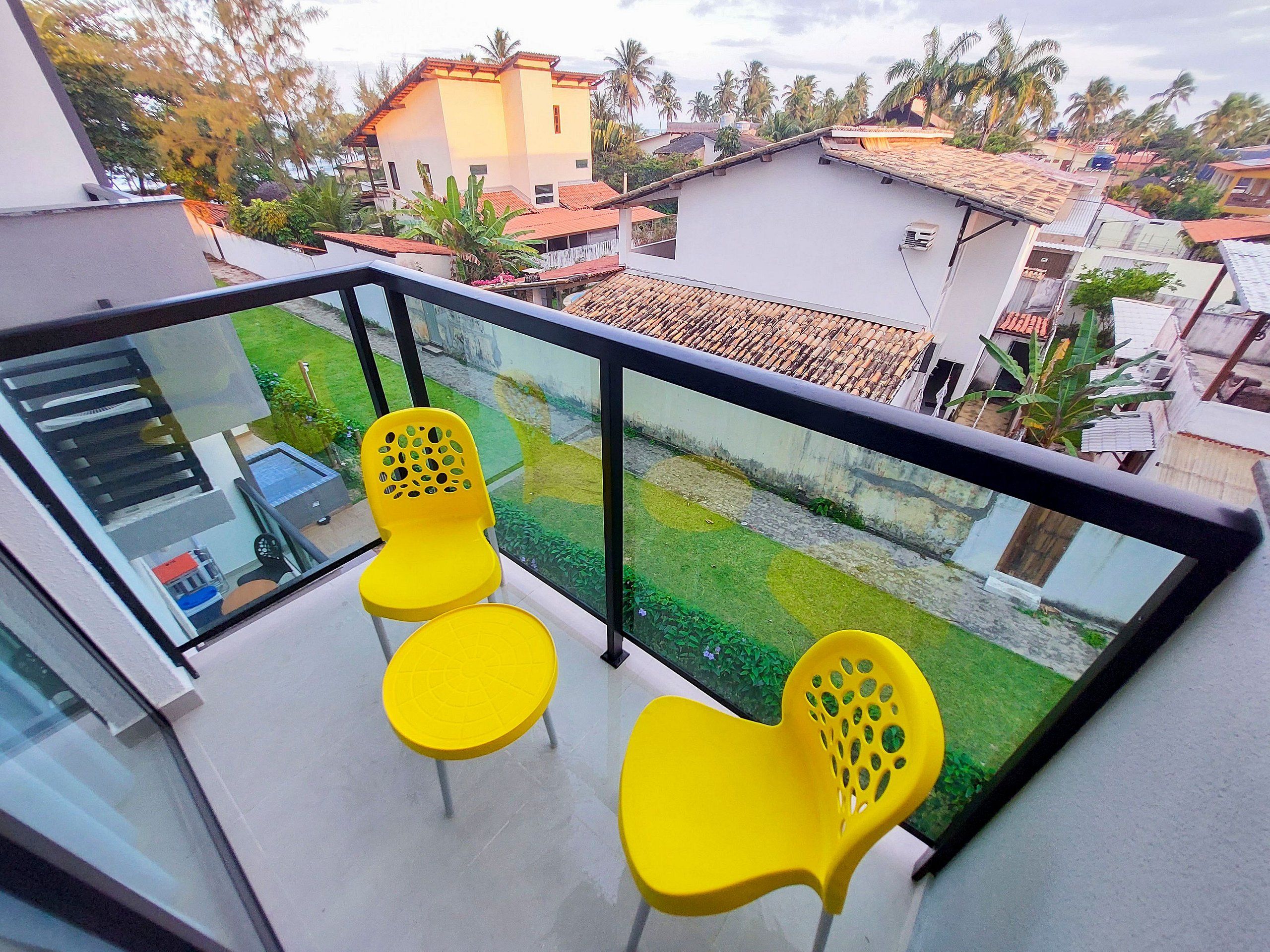 A balcony with yellow chairs, a glass railing, and a view of neighboring houses and greenery.