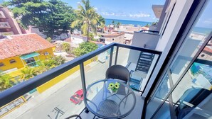 A balcony with a glass table and two chairs, overlooking a street with buildings and a clear sky. - Lindos Flats - Maraca Beach Residence II (Ipojuca)