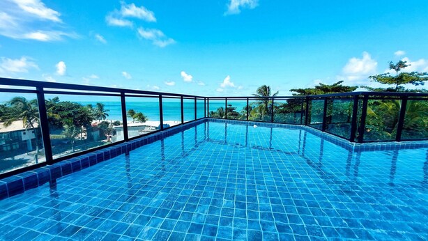A clear blue infinity pool with a glass balustrade, surrounded by lush greenery and a view of the ocean. - Lindos Flats - Maraca Beach Residence II (Ipojuca)