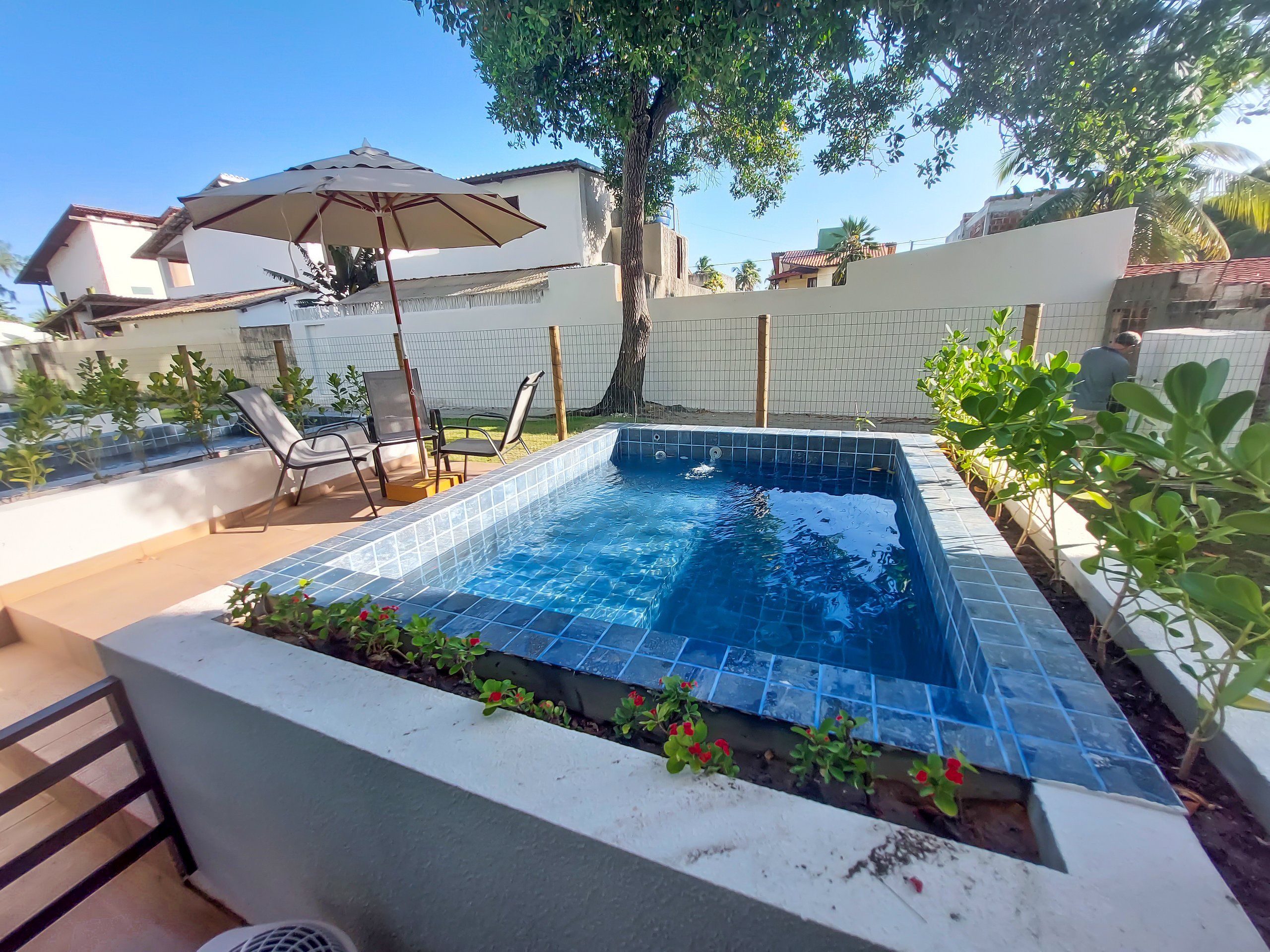 A swimming pool with tiled edges, surrounded by a low wall, and a few chairs and an umbrella.