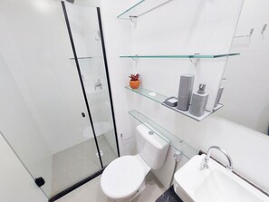 A modern bathroom with a glass shower enclosure, a toilet, a sink with a mirror, and glass shelves with toiletries. - Lindos Flats - Maraca Beach Residence II (Ipojuca)