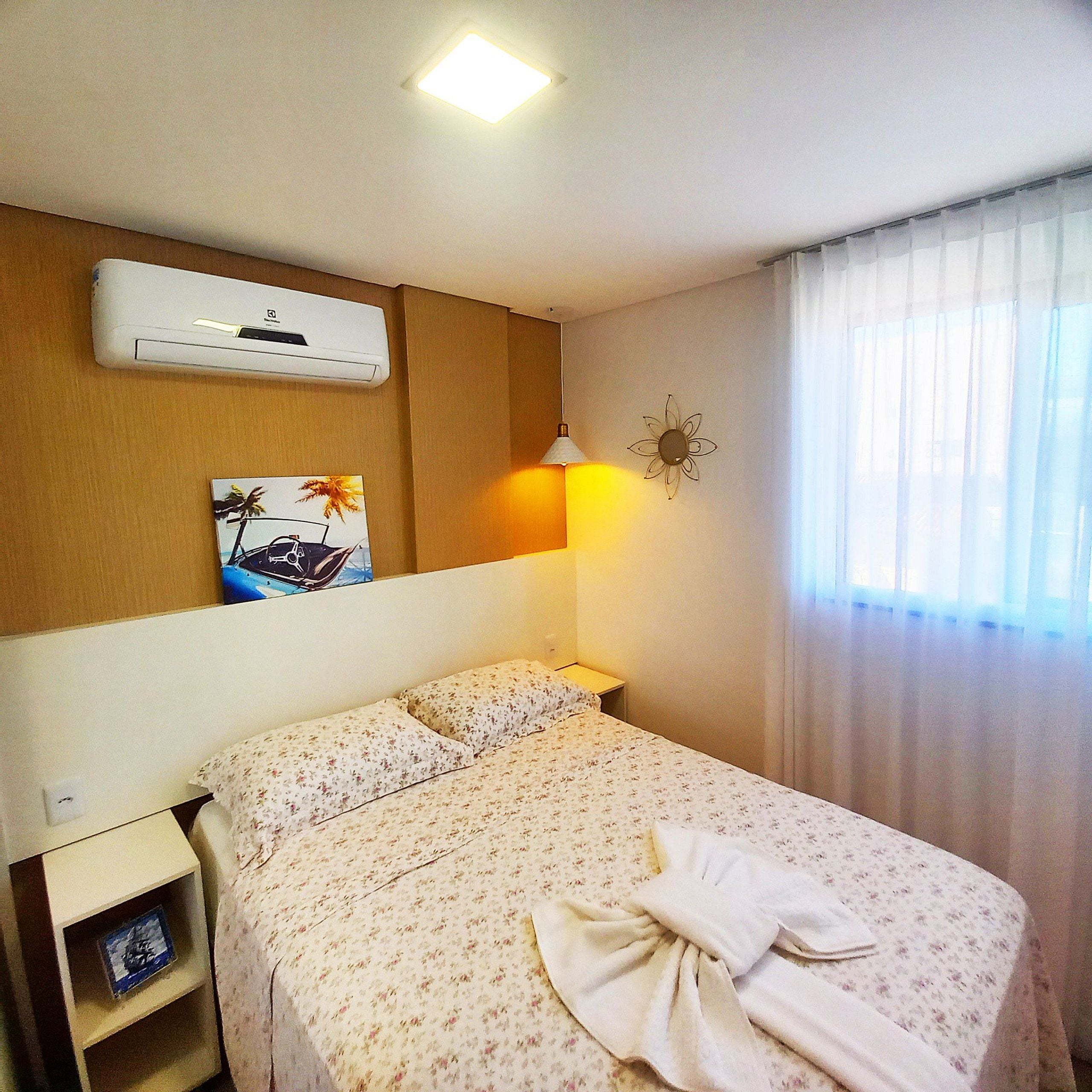 Ein Hotelzimmer mit Bett, Nachttisch, Wandairconditioner, Wanduhr, Wanddeko und einem Vorhangfenster.