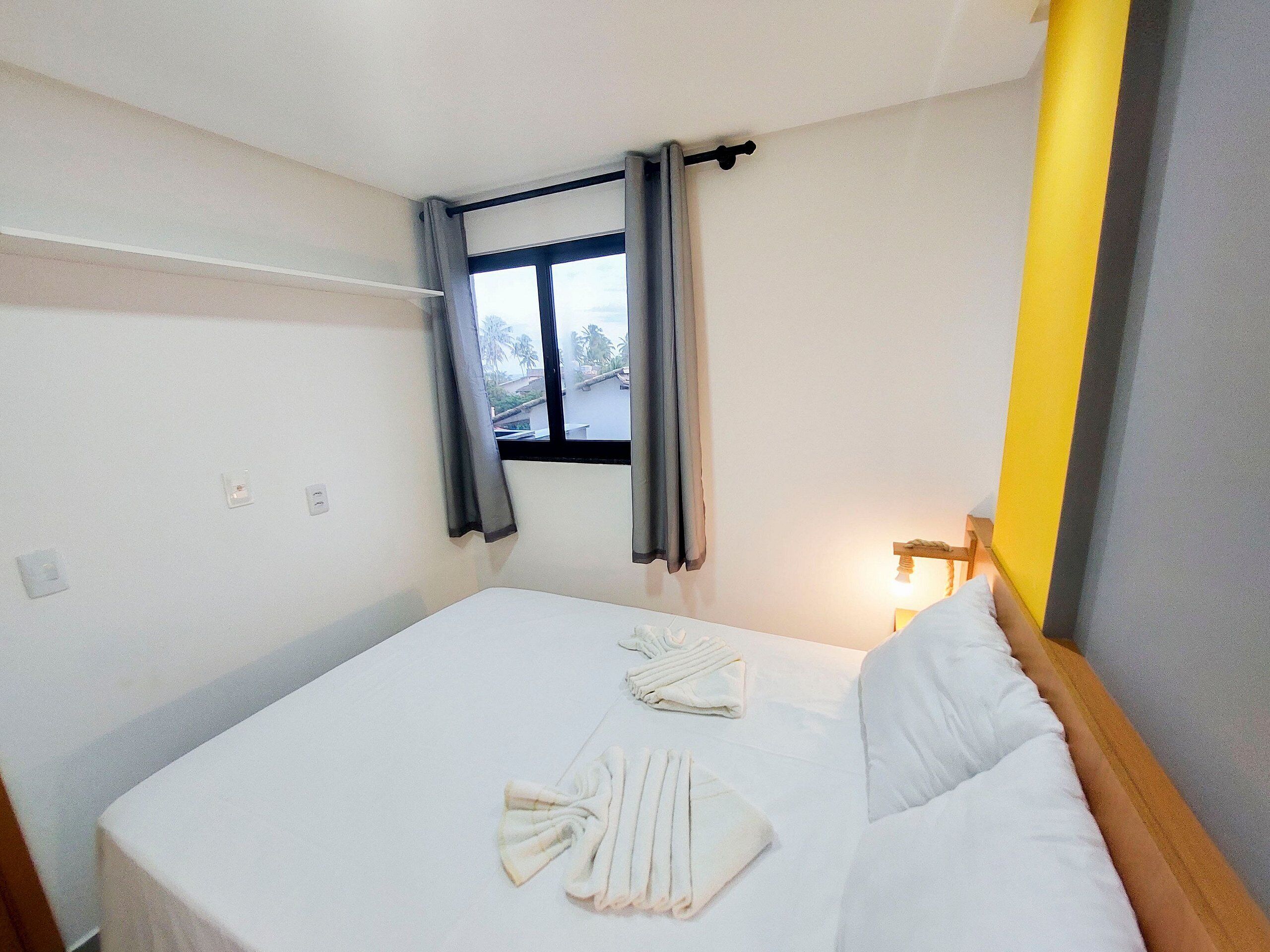 A single bed with white linens, a bedside table with a lamp, and a window with curtains.
