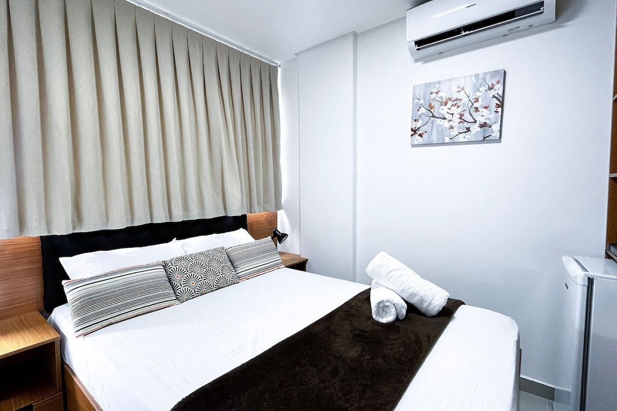 A neatly made bed with a dark brown throw, a bedside lamp, and a rolled towel on the side.