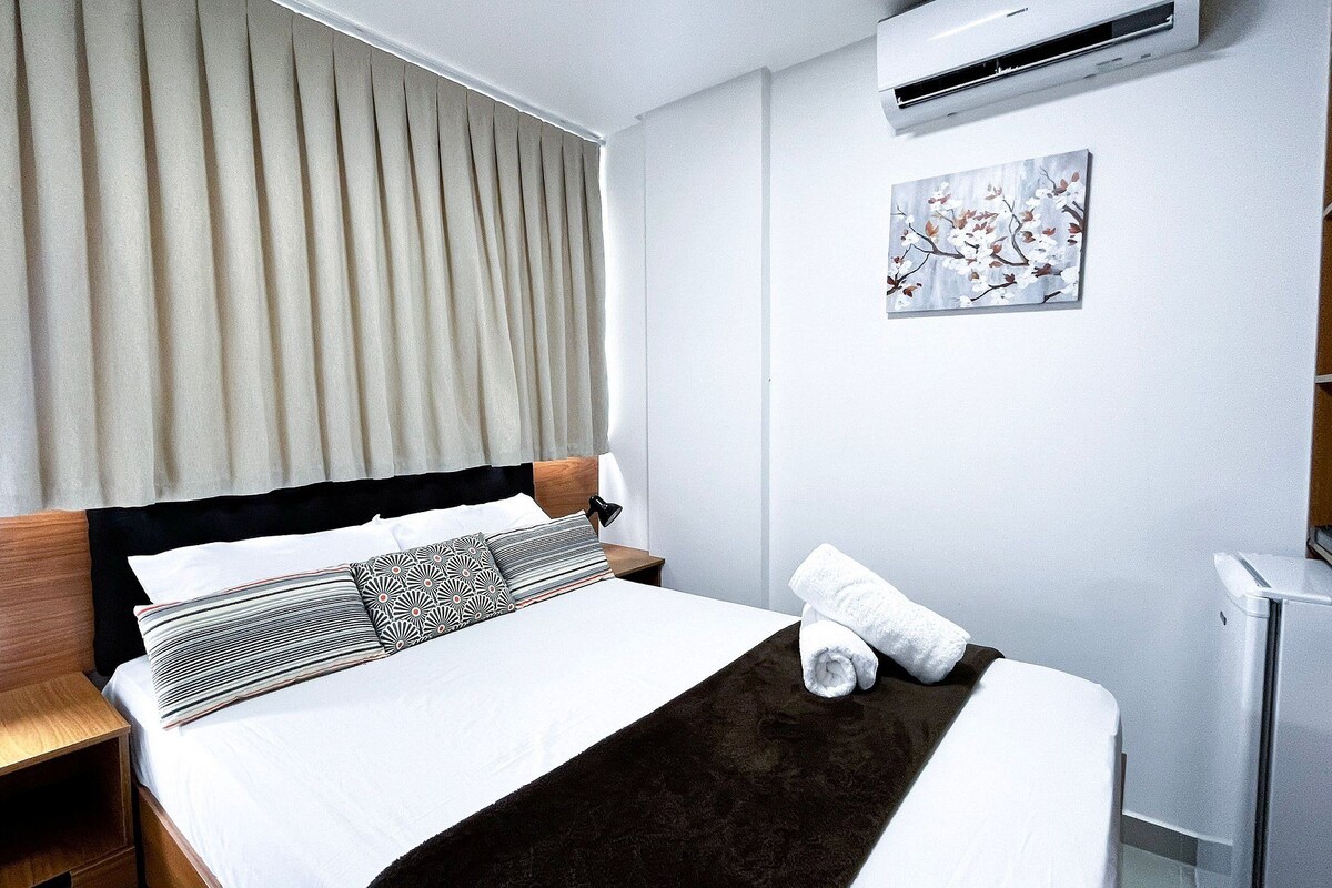 A neatly made bed with a dark brown throw, a bedside lamp, and a rolled towel on the side.