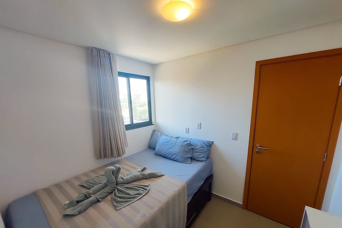 A small, single-bed room with a window, a door, and a light fixture.