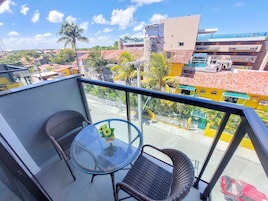 Varanda com duas cadeiras de vime e uma mesa de vidro, com vista para uma área residencial com edifícios coloridos e palmeiras.