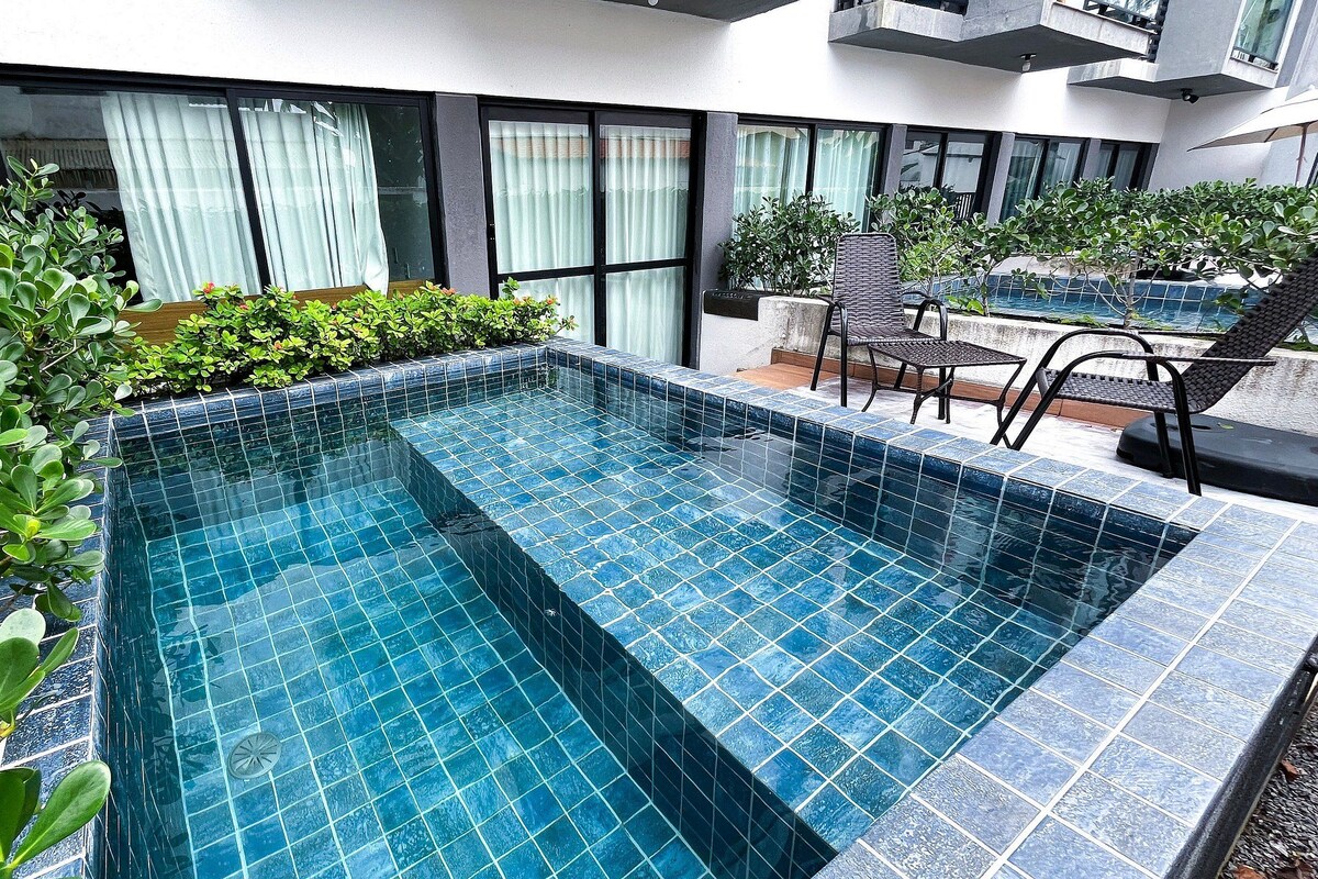 A rectangular outdoor swimming pool with blue tiles, surrounded by greenery and modern outdoor furniture.