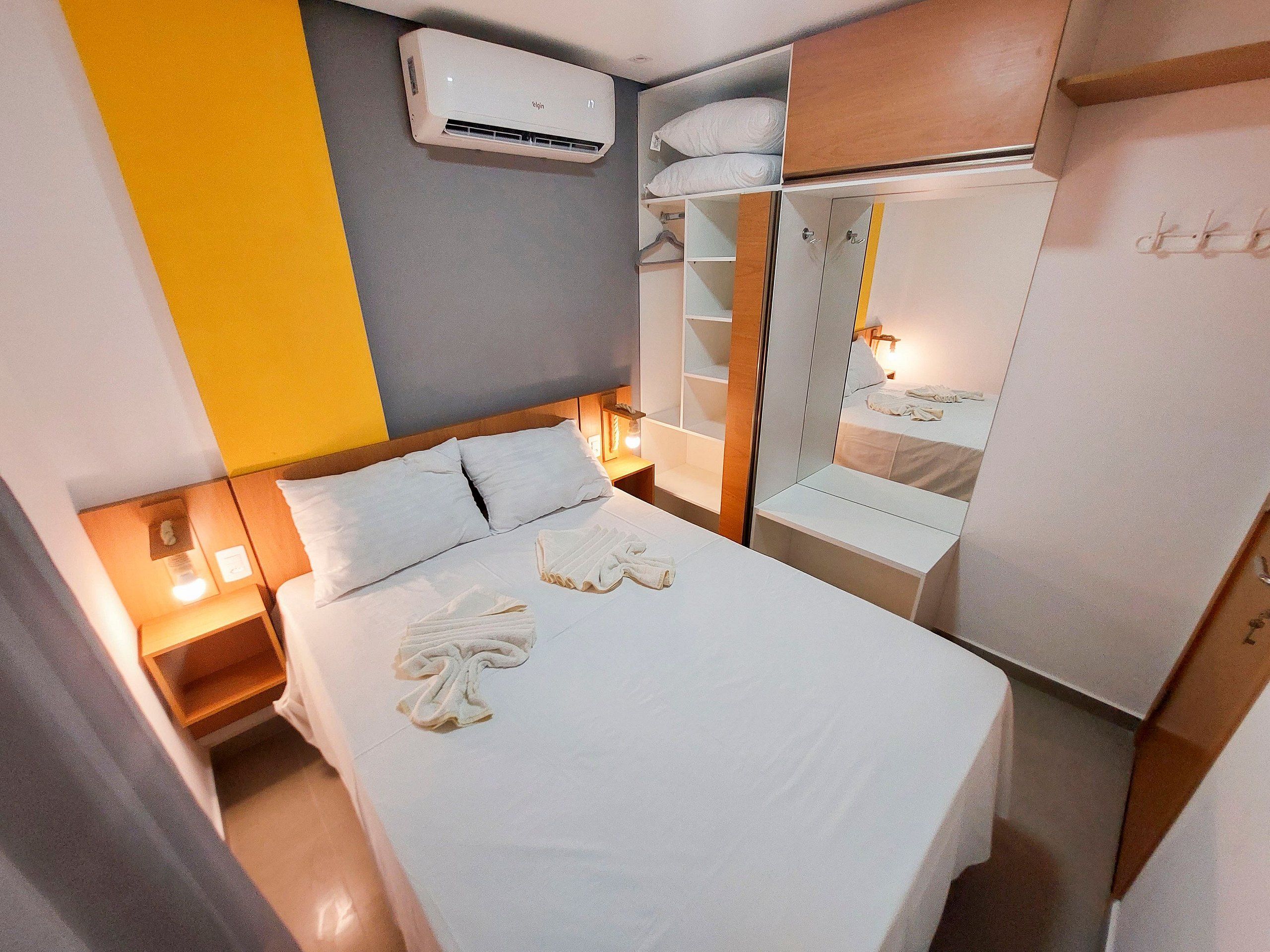 A hotel room with a bed, bedside tables, an air conditioning unit, a wardrobe, and a mirror.