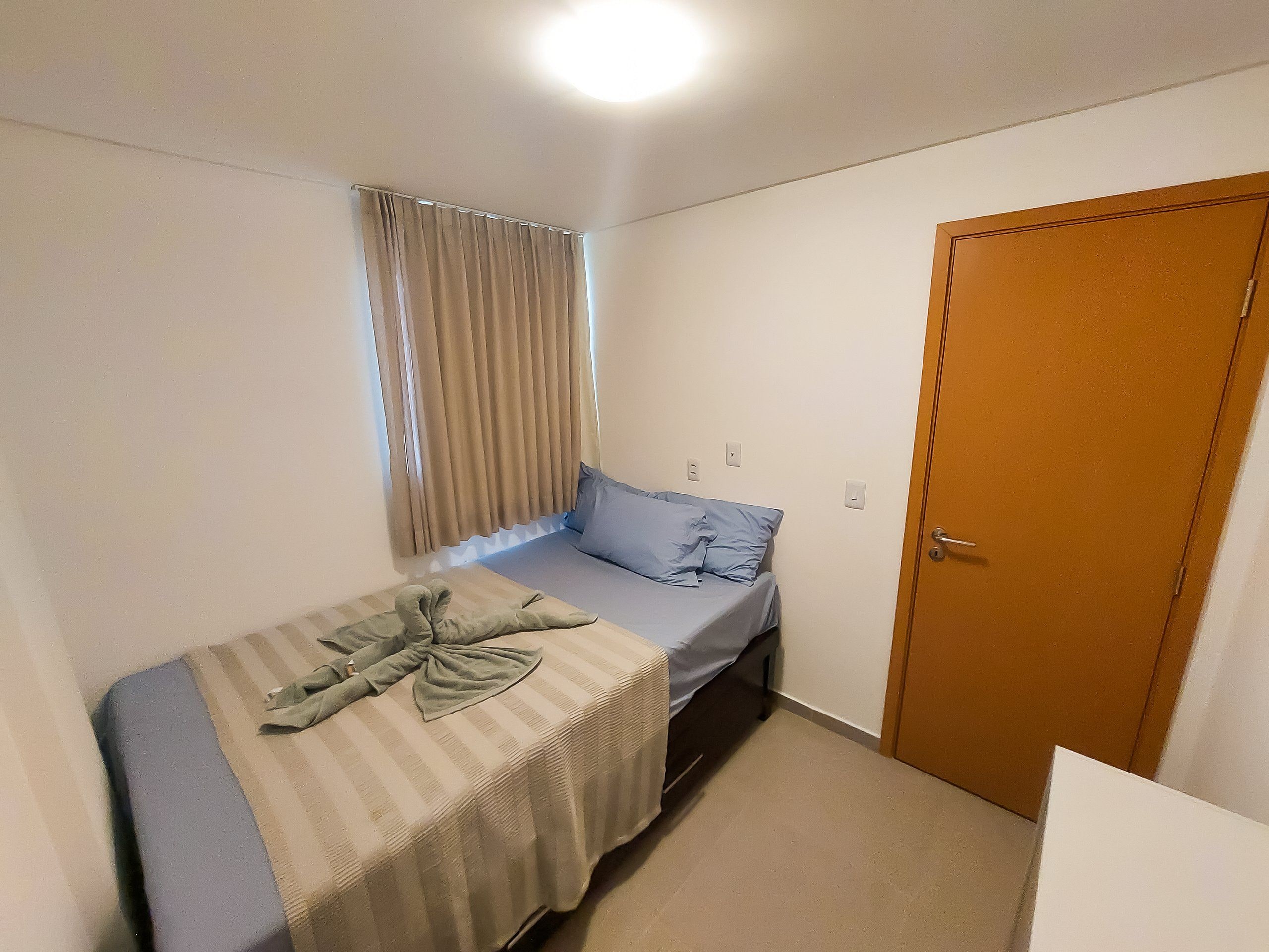 Una habitación pequeña con cama individual, puerta de madera, ventana con cortinas y un interruptor de luz en la pared.