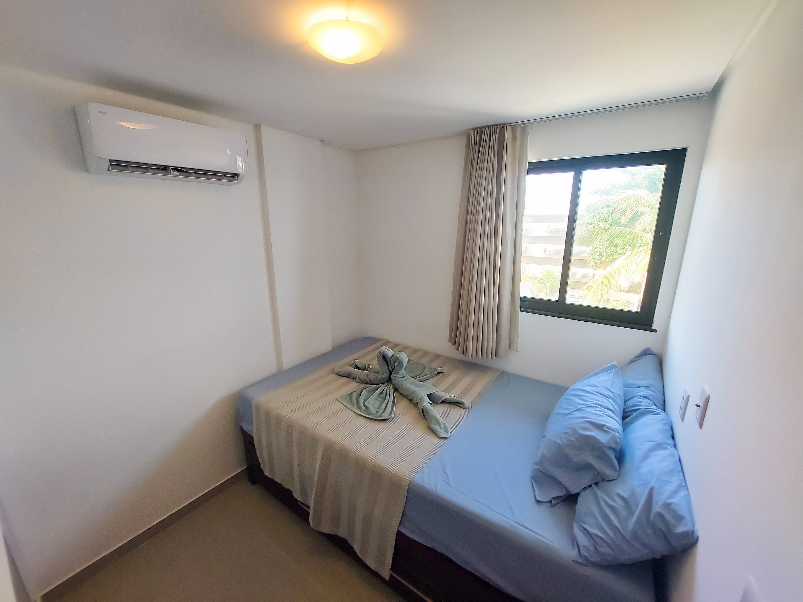Una habitación pequeña con cama individual, ventana, aire acondicionado de pared y luz en el techo.